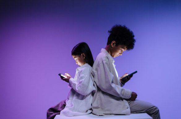 Zwei Kinder unterschiedlichen Alters sitzen Rücken an Rücken und benutzen Smartphones.
